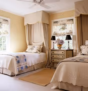 Sunlit bedroom with two twin beds, floral window shades, beige wallpaper, and a shared bedside table with lamps.
