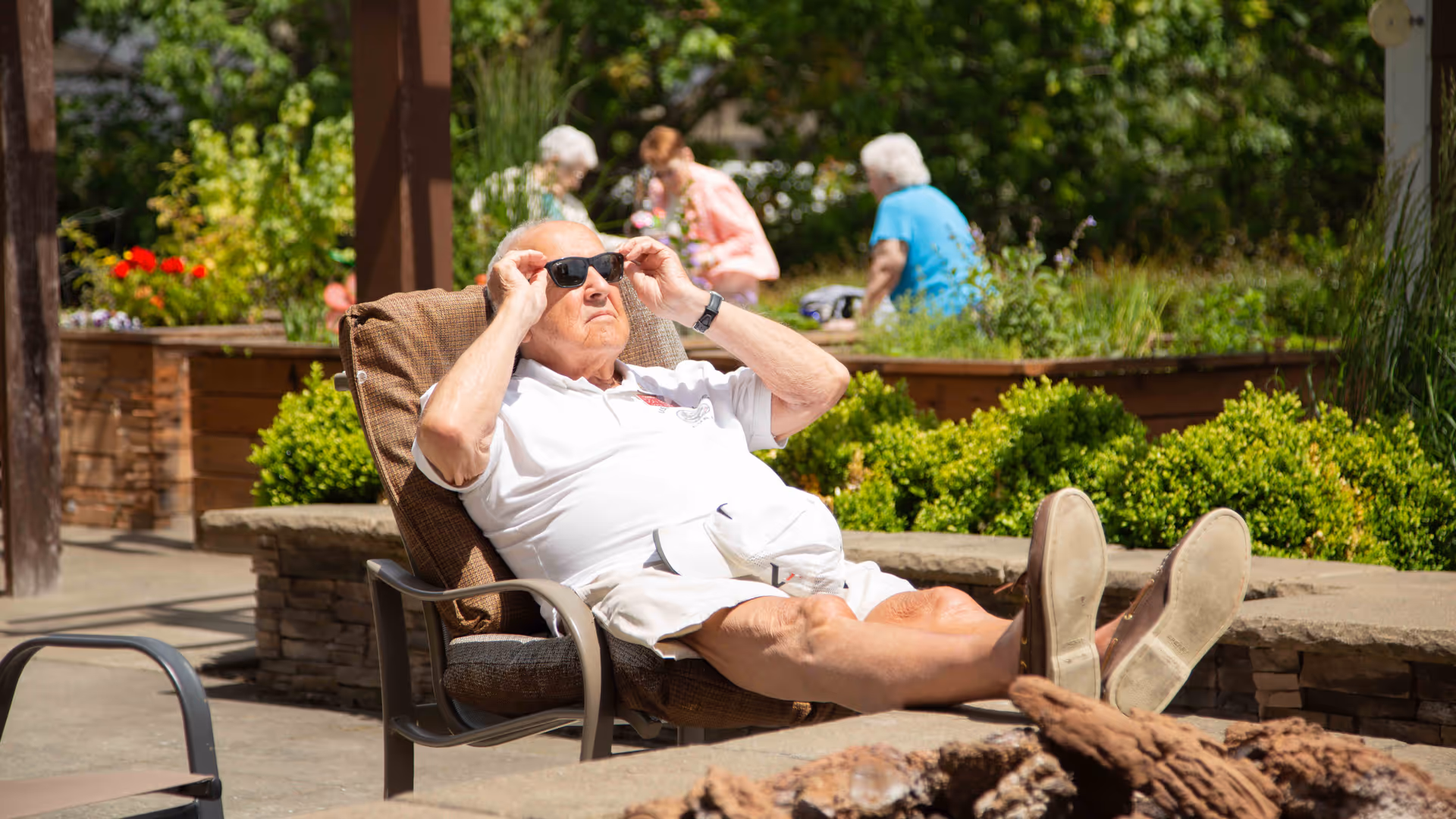 An elderly man wearing sunglasses and a white outfit is reclining comfortably on a cushioned outdoor chair with his feet up, enjoying a sunny day in a garden area. In the background, three elderly women are engaged in conversation near raised garden beds filled with greenery and flowers.