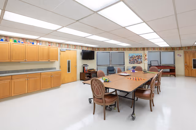 A bright, spacious activity room with a large rectangular table in the center surrounded by chairs. The table has board games and craft supplies on it. The room features wooden cabinets along one wall, a wall-mounted TV, and several windows with blinds. The floor is white and the ceiling has fluorescent lighting panels.