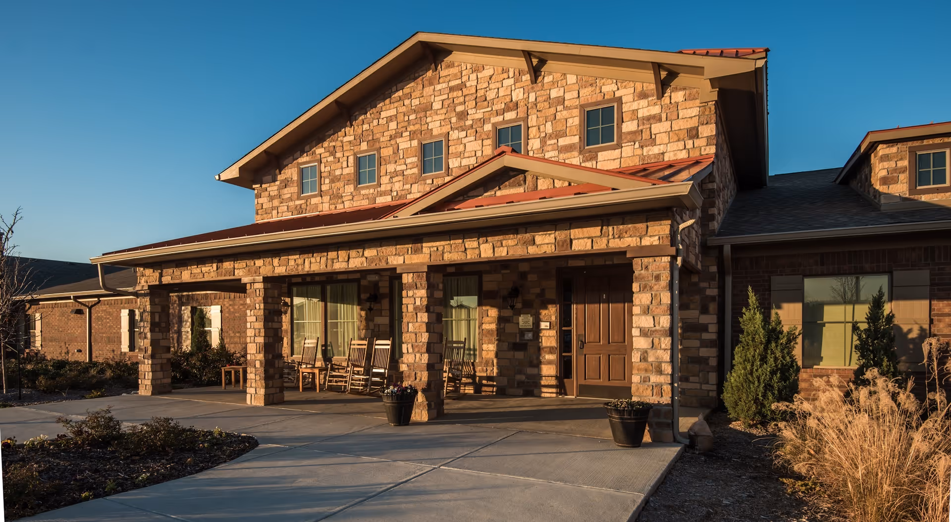 Exterior front view of a stone and brick assisted living facility building with a covered porch featuring rocking chairs and potted plants, under a clear blue sky.