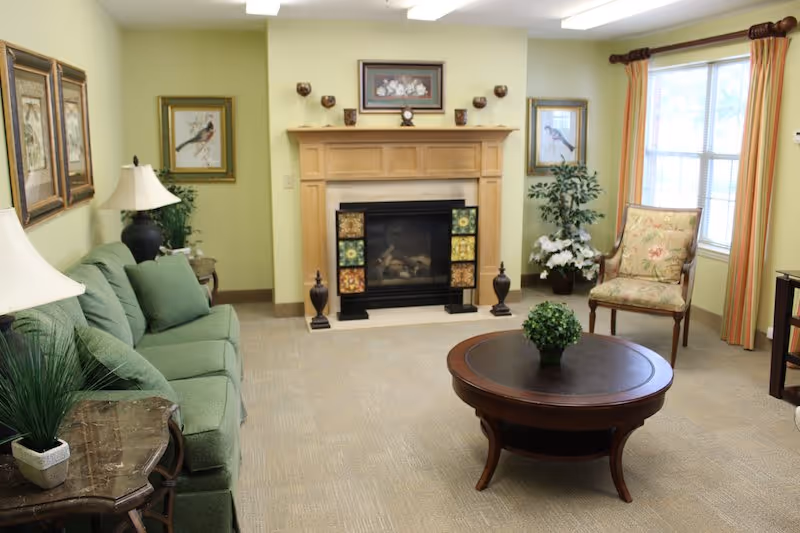 A cozy living room with a green sofa, a wooden coffee table with a small plant, a fireplace with decorative tiles, framed artwork on the walls, a floral armchair near a window with striped curtains, and various plants and lamps.