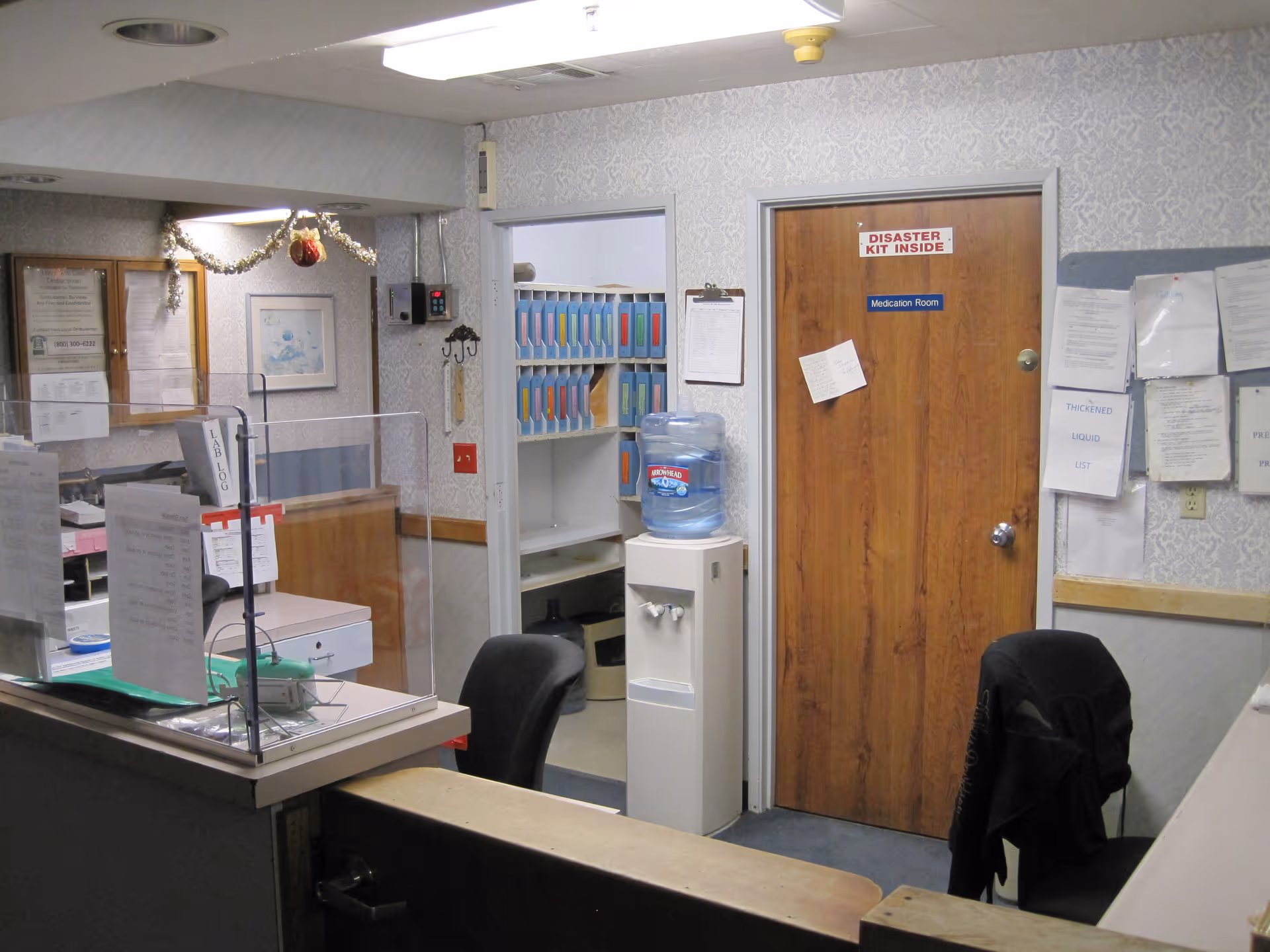 Reception area with a desk featuring a clear protective barrier, office chair, water cooler, and a door labeled 'Medication Room' with a sign above it stating 'Disaster Kit Inside'. The walls have various papers and notices pinned up, and there is a small open doorway showing shelves with organized files.