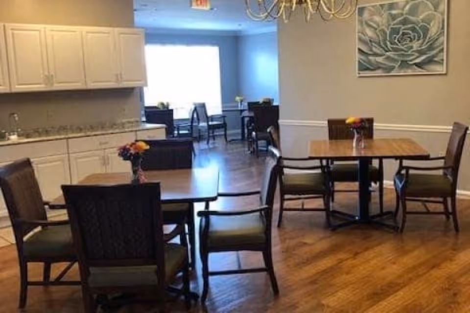 A bright communal dining area with several wooden tables and chairs, a kitchenette with white cabinets, and wall artwork.