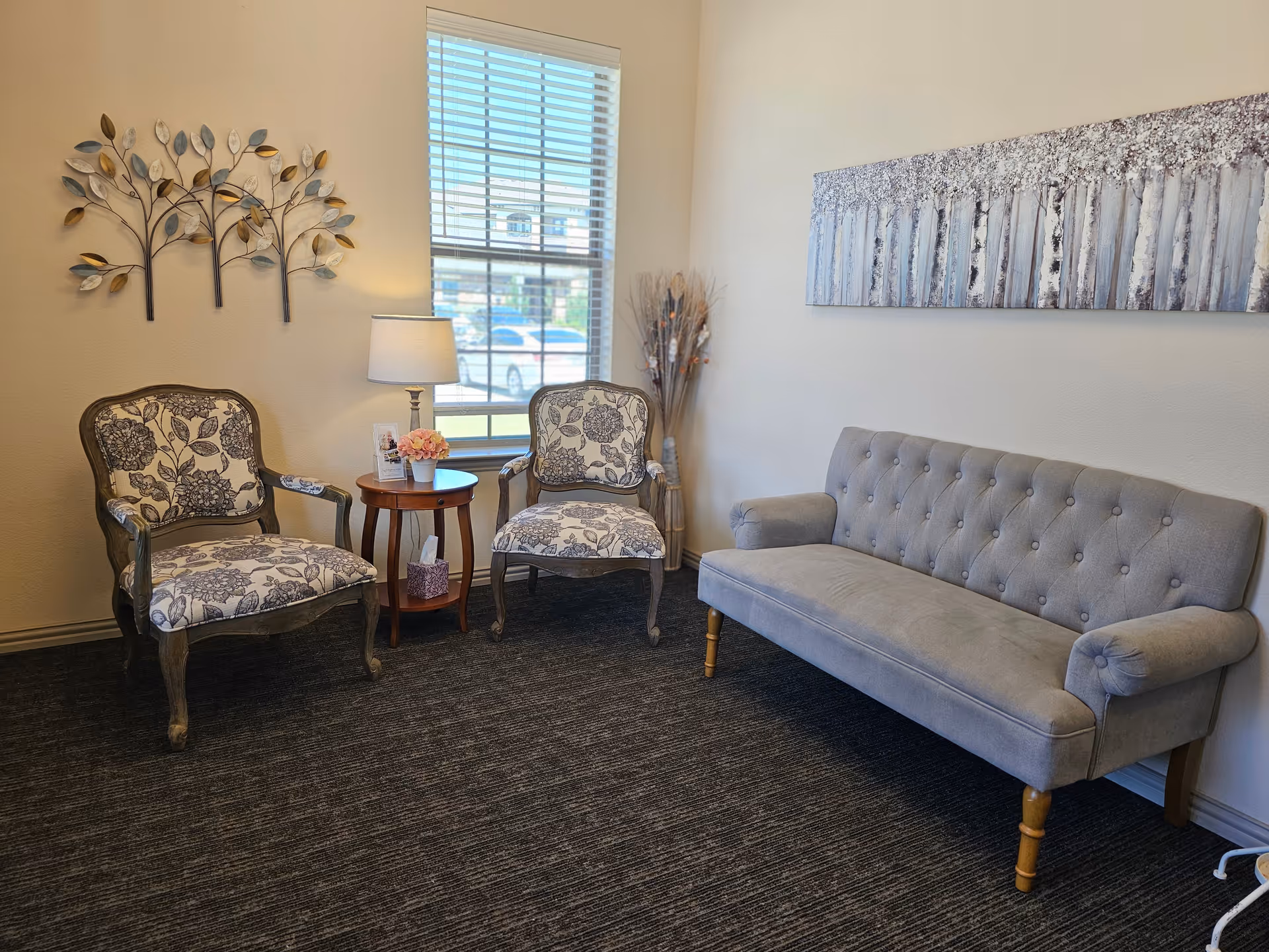 A cozy sitting area with two floral patterned armchairs and a gray tufted sofa. Between the armchairs is a small round wooden table with a lamp, a small flower arrangement, and a tissue box. A window with blinds lets in natural light, and the walls are decorated with a metal leaf wall art and a large painting of birch trees.