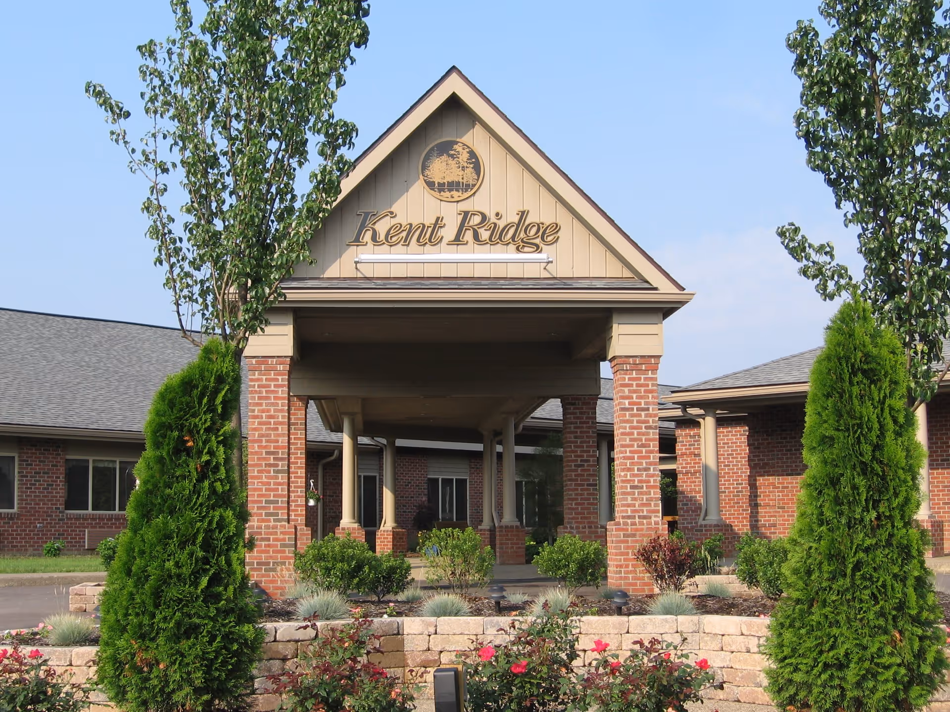 Entrance of The Kent Ridge Senior Living facility with a covered driveway supported by brick pillars, surrounded by landscaped greenery including bushes, flowers, and trees under a clear blue sky.