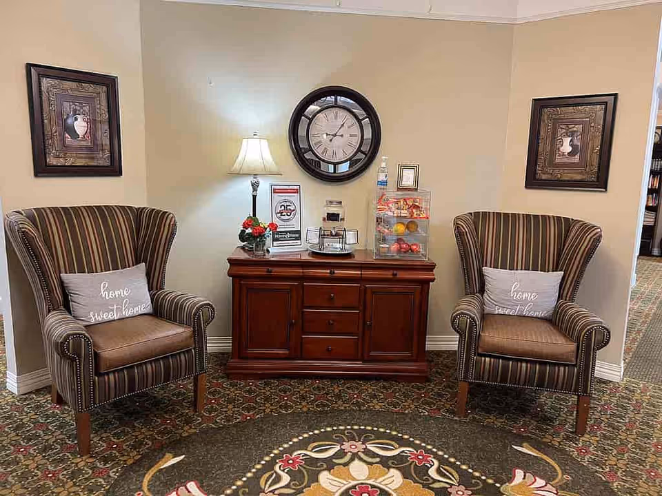 A cozy seating area with two striped armchairs, each with a pillow that says 'home sweet home'. Between the chairs is a wooden cabinet with a table lamp, a clock on the wall above it, framed pictures on either side, and a clear container holding fruit and snacks.