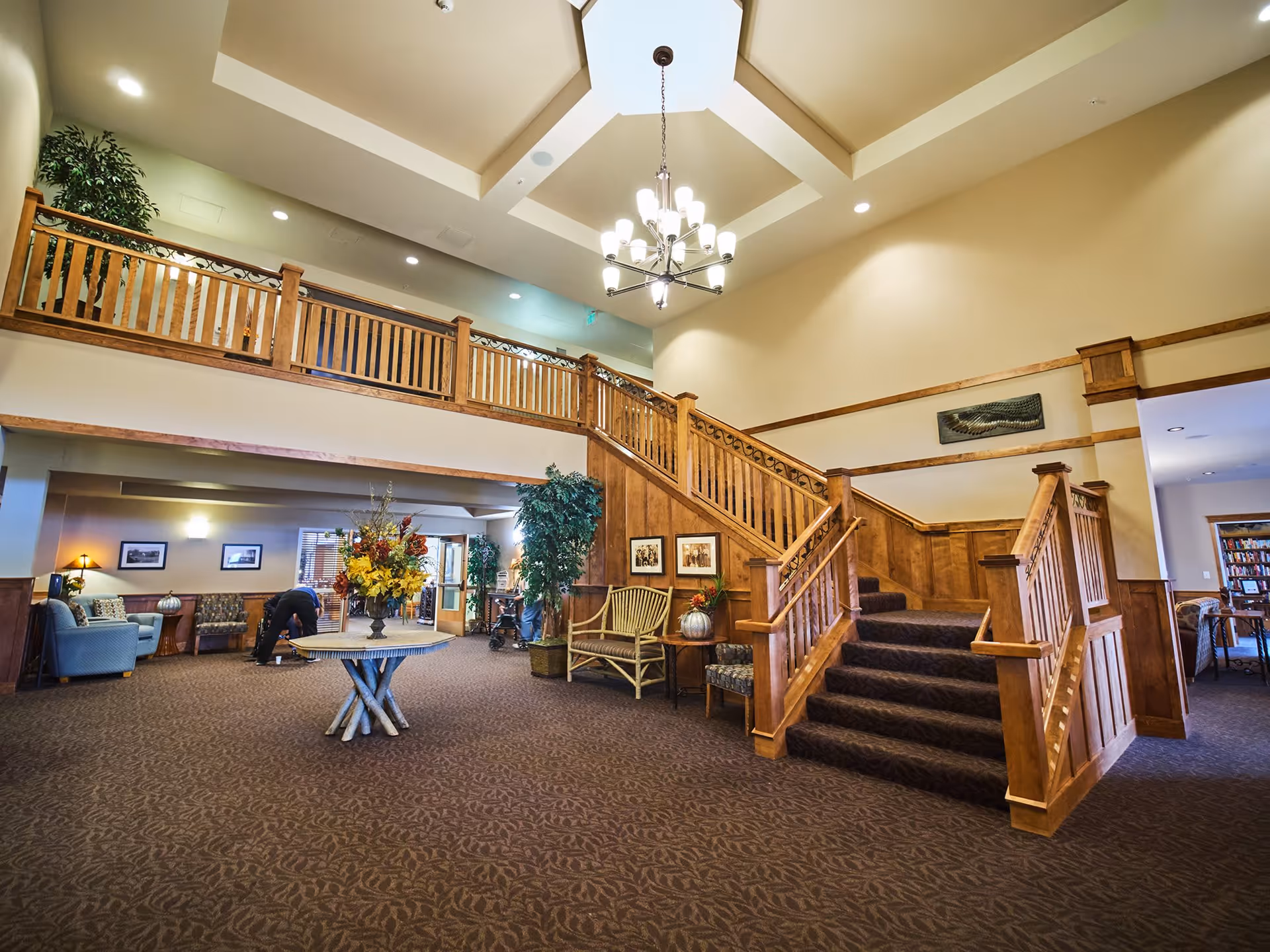 Spacious interior lobby area of Bozeman Lodge featuring a large wooden staircase with carpeted steps, a round table with a floral arrangement in the center, comfortable seating areas with chairs and a bench, decorative plants, framed pictures on the walls, and a chandelier hanging from a high ceiling with a skylight.