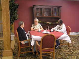 Three elderly women sitting around a table with a red tablecloth in a room with a burgundy wall and a wooden cabinet behind them. They appear to be engaged in conversation or enjoying a meal together.
