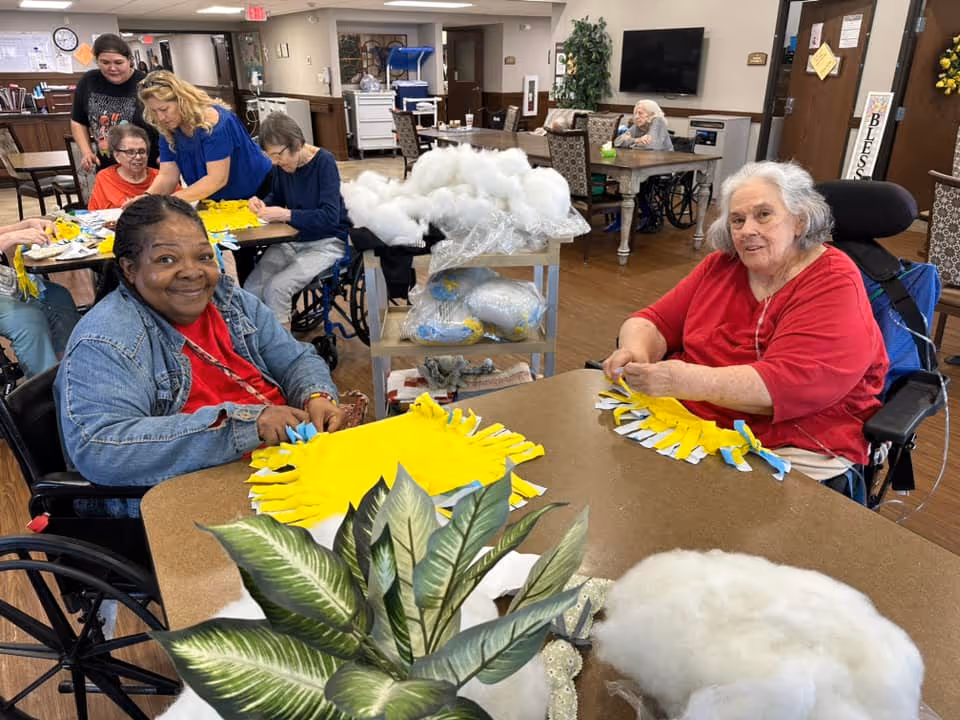 A group of elderly women, some in wheelchairs, are seated around tables in a communal room engaging in a craft activity with yellow fabric. The room has wooden floors, tables, chairs, and a TV mounted on the wall. There are plants and craft materials visible, and a staff member is assisting one of the women.