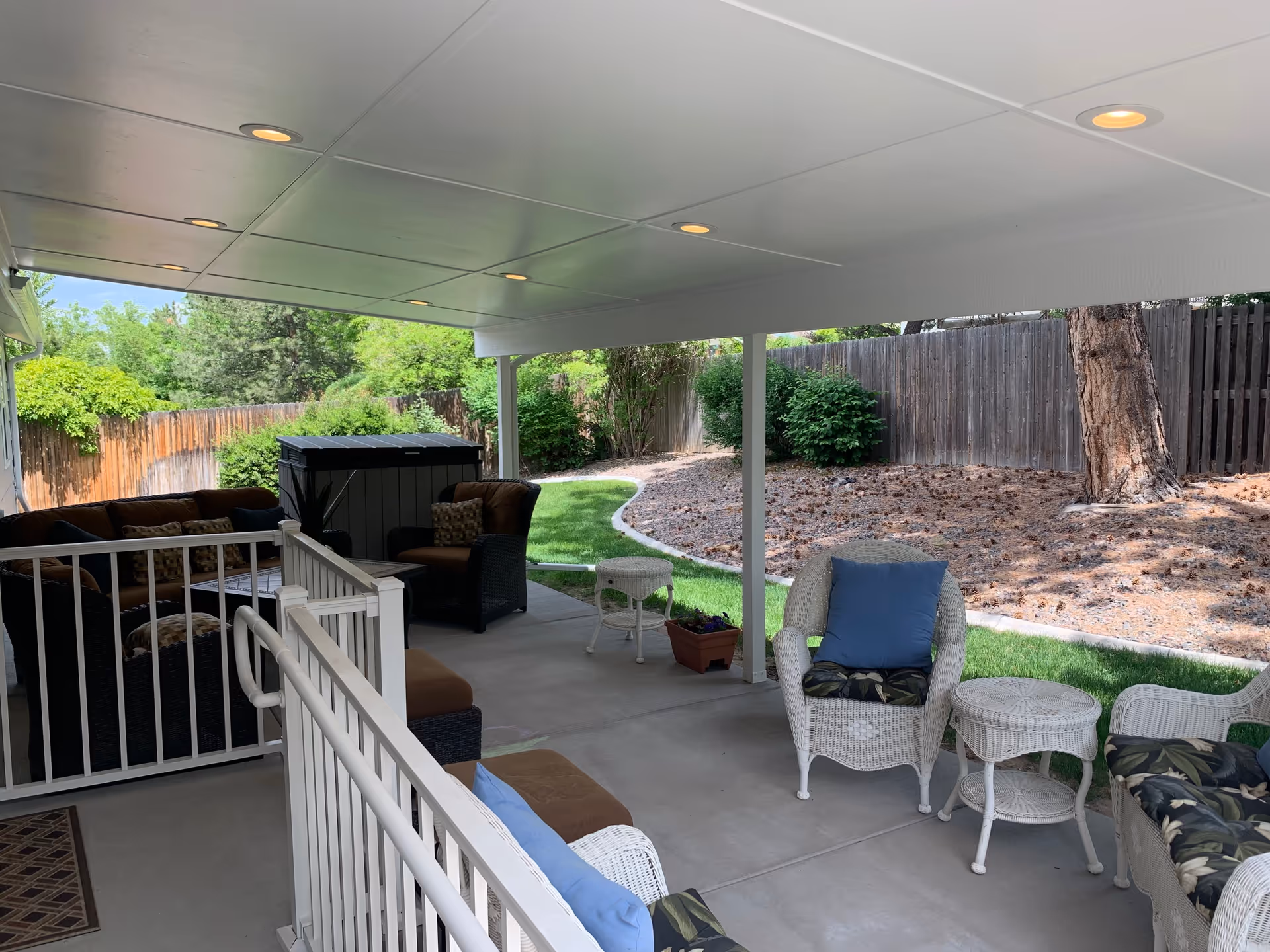 Covered outdoor patio area with white wicker chairs and tables, cushioned seating, and a brown cushioned sofa set. The patio overlooks a fenced backyard with green grass, bushes, and trees under a clear sky.