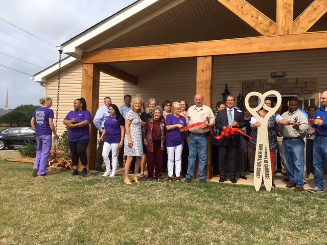 A group of people standing in front of a building entrance for a ribbon-cutting ceremony, holding a large decorative pair of scissors and a red ribbon.