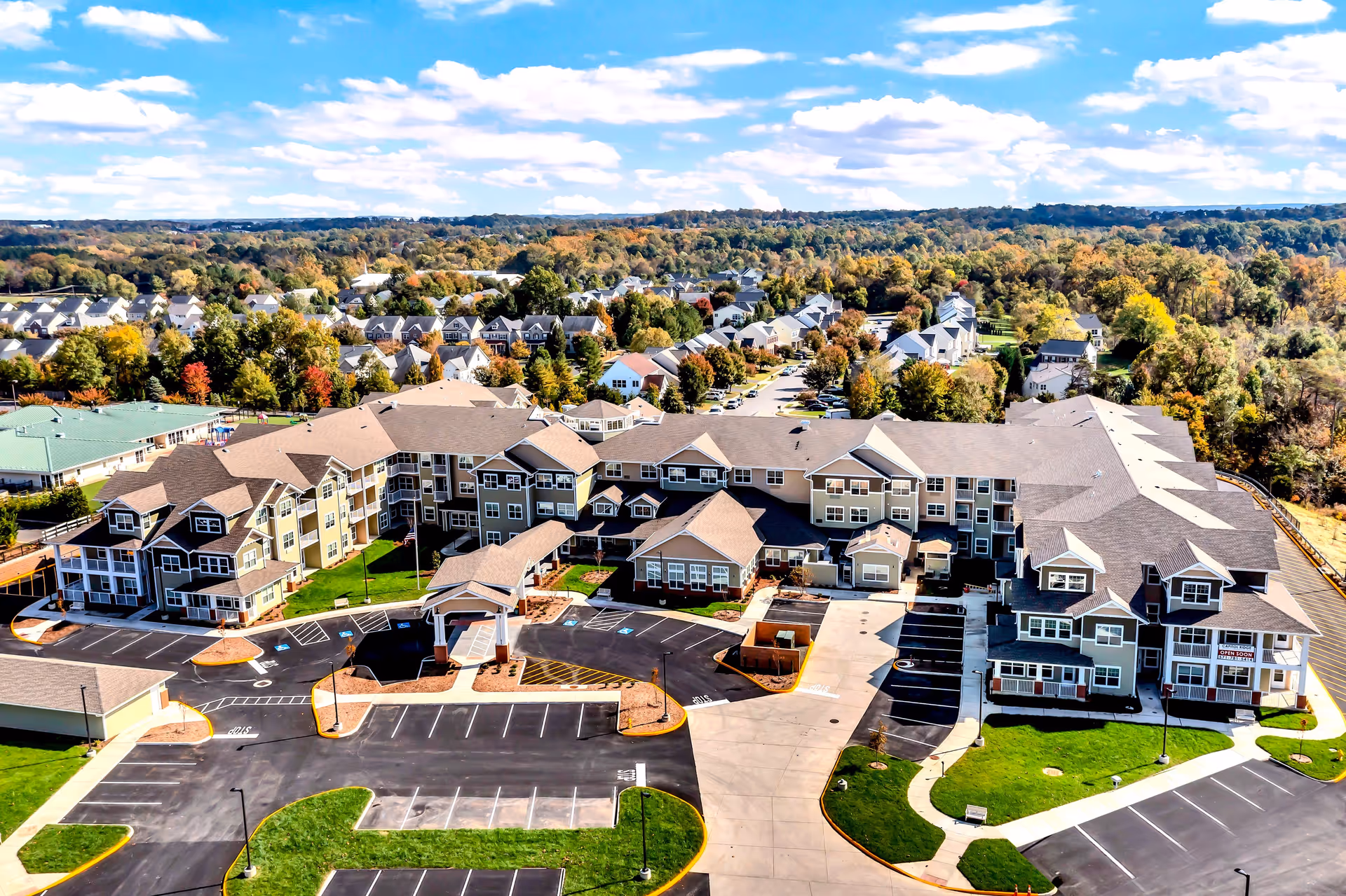 Aerial view of Capitol Ridge Gracious Retirement Living facility showing a large multi-wing building with multiple floors, surrounded by parking lots and green lawns. The background includes a suburban neighborhood with many houses and trees under a partly cloudy blue sky.