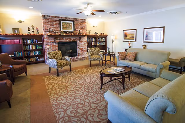 A cozy living room with a patterned carpet, two beige sofas, two patterned armchairs, a wooden coffee table with magazines, and a brick fireplace flanked by bookshelves. The room is softly lit with wall sconces and a ceiling fan with lights. There are framed pictures on the walls and decorative items on the mantel and shelves.