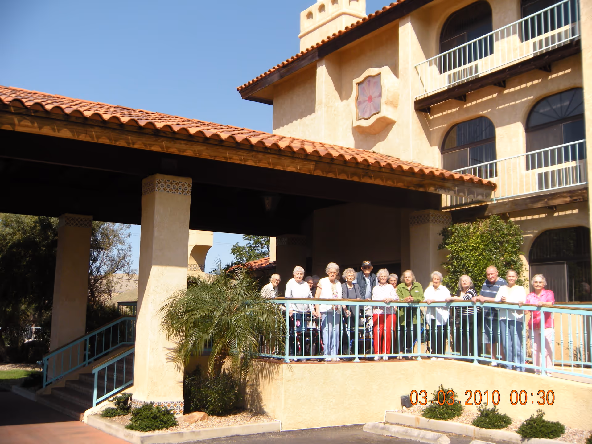 A group of elderly people standing together on a balcony outside a beige stucco building with a red tile roof under a clear blue sky.
