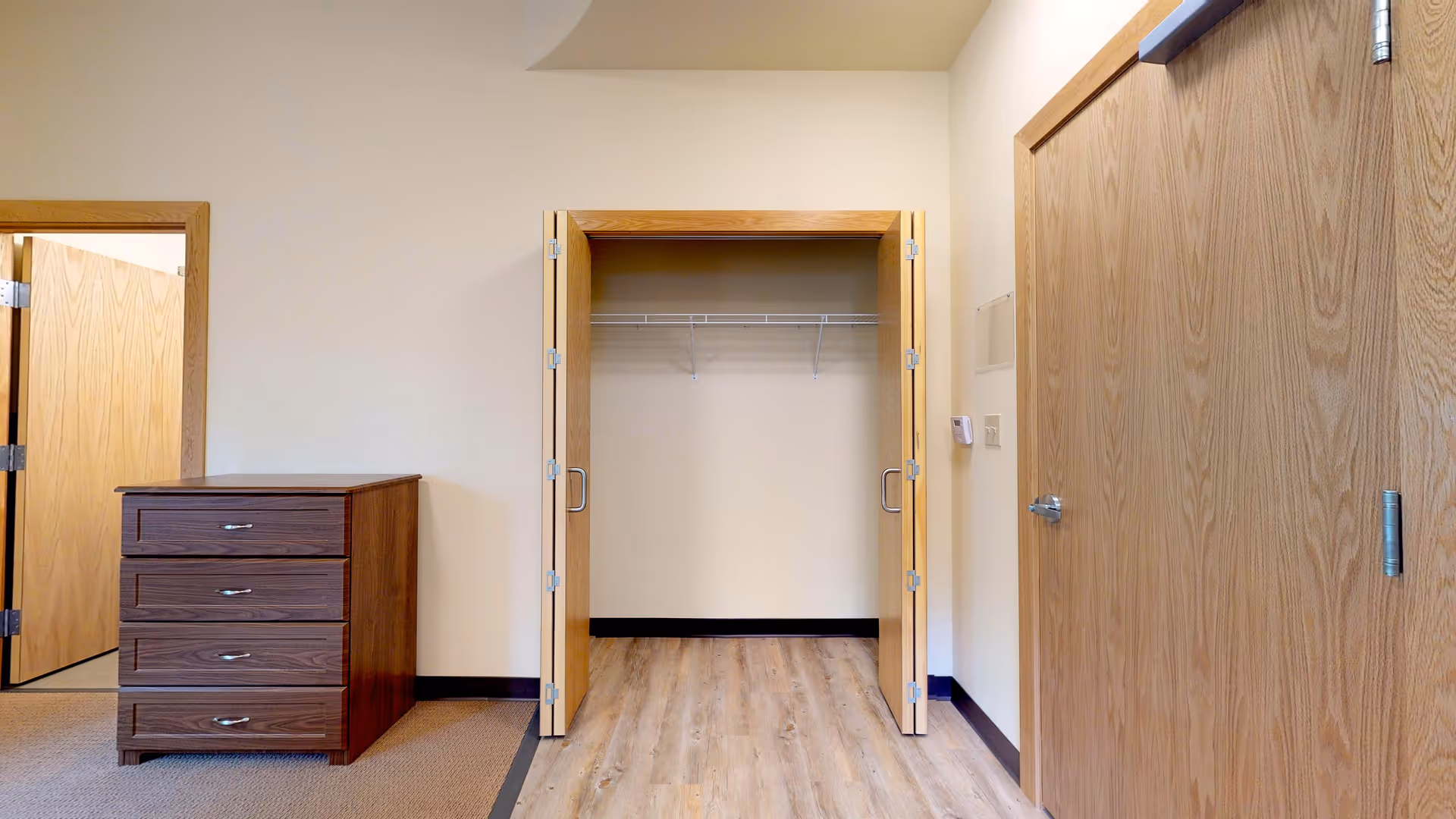 An empty room with light beige walls and wood flooring. There is an open closet with double wooden doors and a white wire shelf inside. To the left of the closet is a wooden chest of drawers with four drawers. There are two closed wooden doors visible, one on the left side and one on the right side of the image.