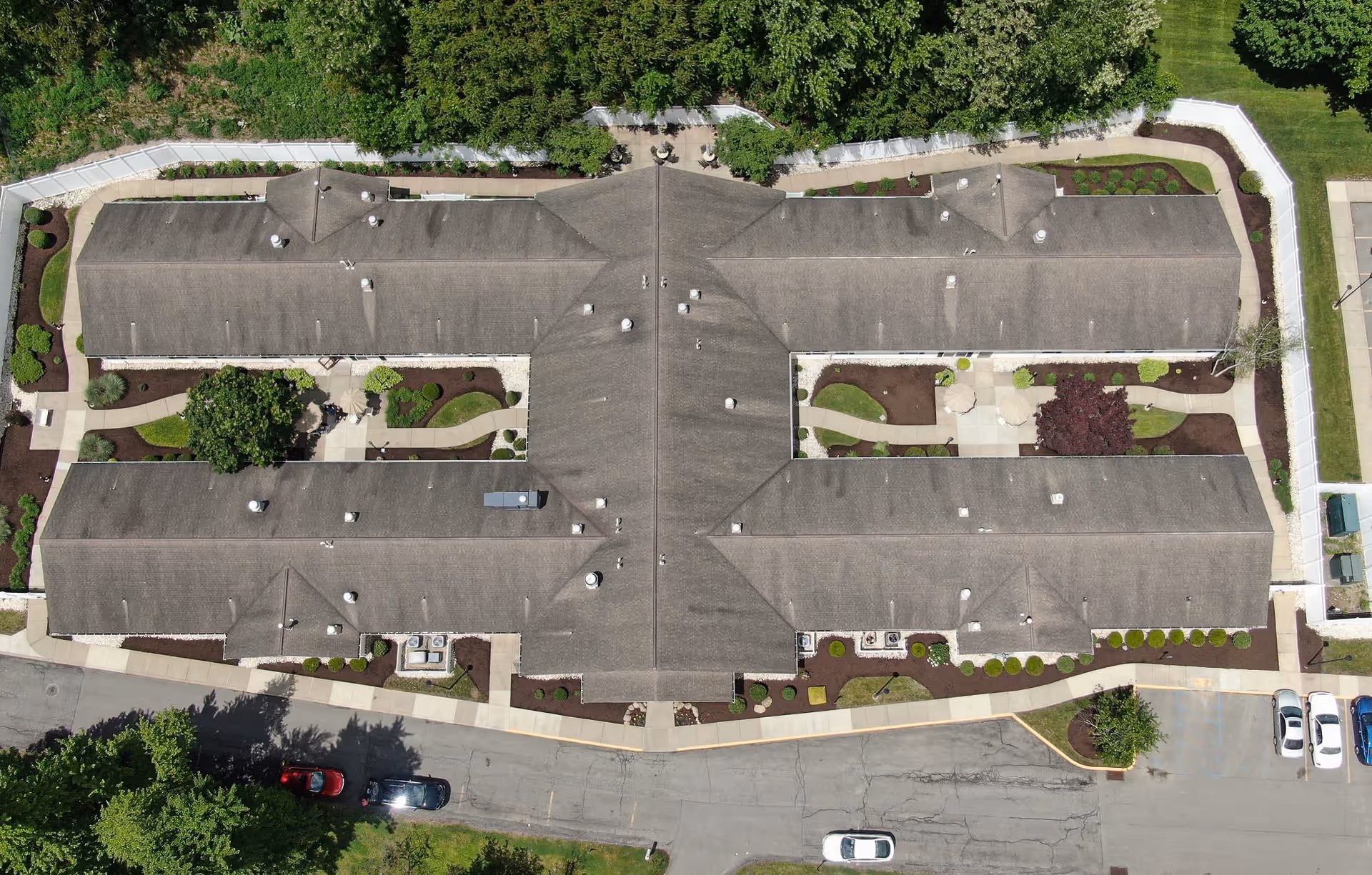 Aerial view of a large, single-story building with a complex roof structure surrounded by landscaped gardens, walkways, and parking areas. The building is enclosed by a white fence and bordered by trees and greenery.