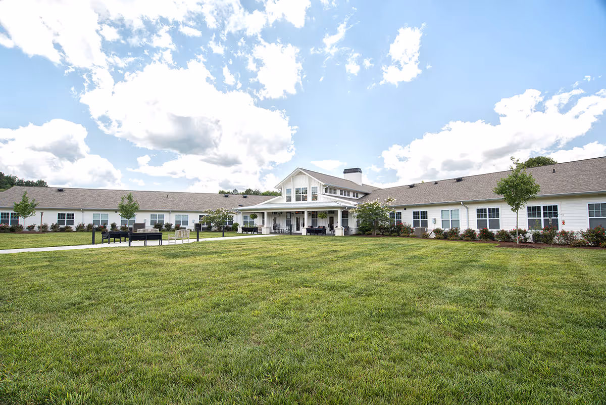 Wide exterior view of Dominion Senior Living of Athens building with a large green lawn in the foreground, a paved walkway leading to the entrance, and a partly cloudy blue sky above.