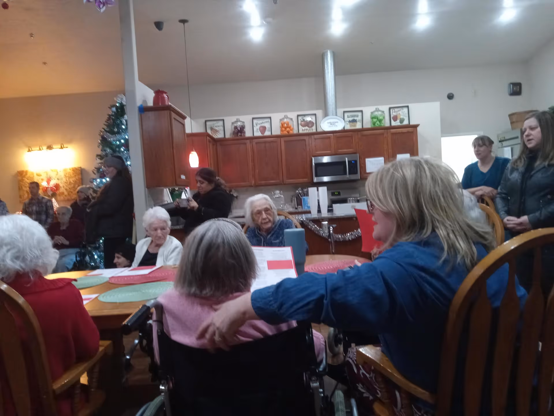 A group of elderly people and caregivers gathered in a communal dining area with wooden chairs and tables. The background shows a kitchen with wooden cabinets, a microwave, and decorative jars on top of the cabinets. A decorated Christmas tree is visible on the left side, indicating a festive atmosphere.