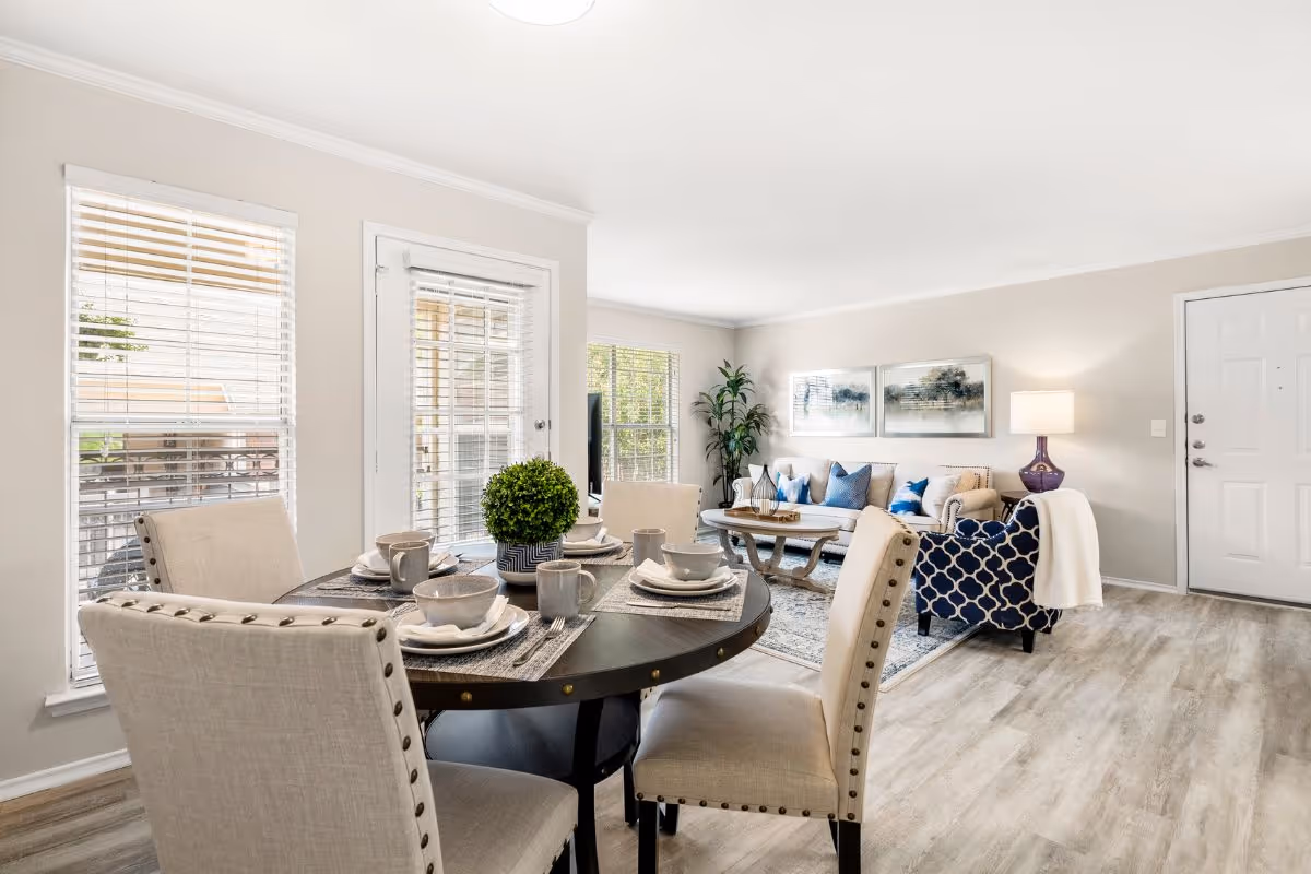 Bright and modern living and dining area with a round dining table set for four, beige upholstered chairs, a beige sofa with blue and white pillows, a patterned armchair, a coffee table, a floor lamp, and large windows with blinds letting in natural light.