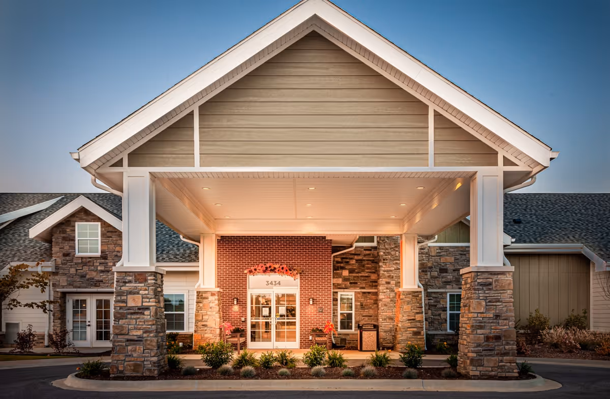 Front entrance of a senior living facility with a covered drop-off area supported by stone pillars. The building features a mix of brick and stone exterior with large windows and a peaked roof. There are plants and shrubs lining the entrance walkway.