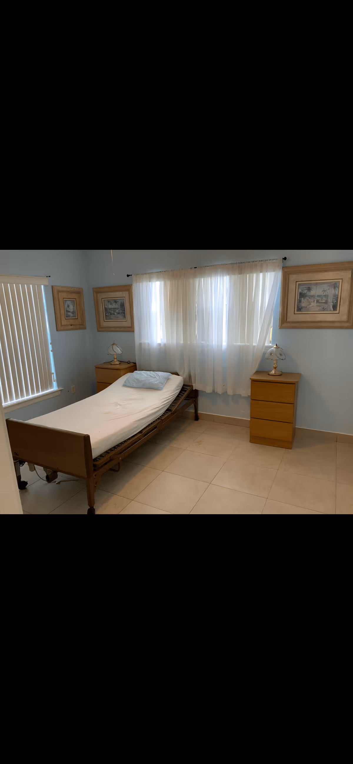 A simple bedroom with a single hospital-style bed with a white sheet and a blue pillow. The room has light blue walls, two wooden nightstands with lamps on each side of the bed, and three framed pictures hanging on the walls. There are sheer white curtains covering a window behind the bed and vertical blinds on a window to the left.