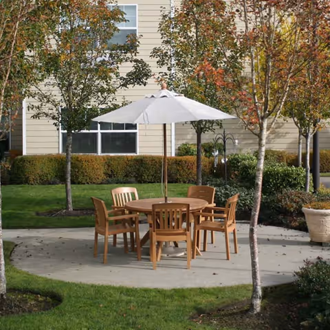 Outdoor patio area with a round wooden table and six matching chairs under a large white umbrella, surrounded by trees and greenery in front of a beige building.