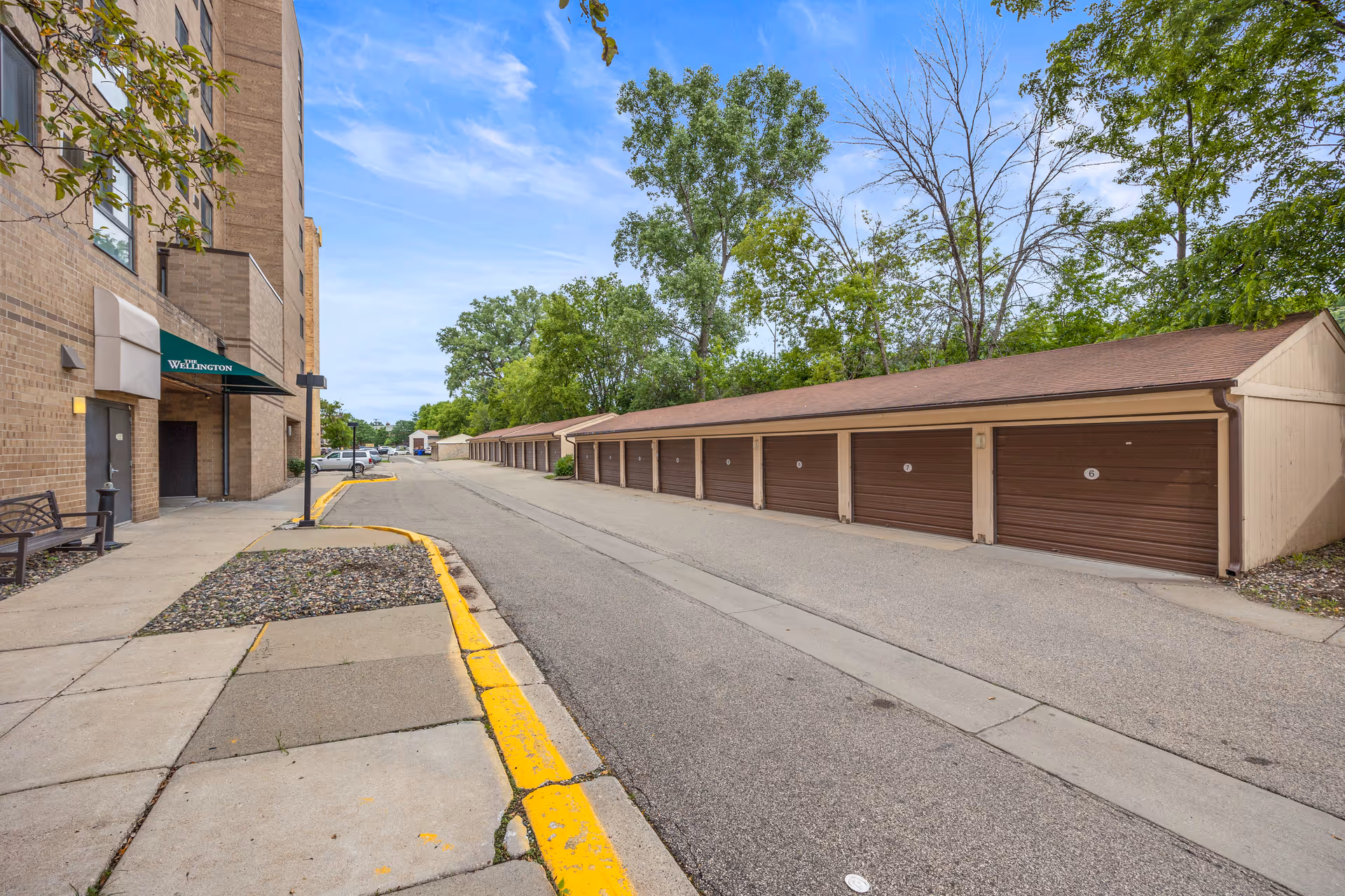View of a paved driveway lined with a row of brown garage doors on the right and a beige brick building with a green awning labeled 'The Wellington' on the left, surrounded by trees under a partly cloudy sky.