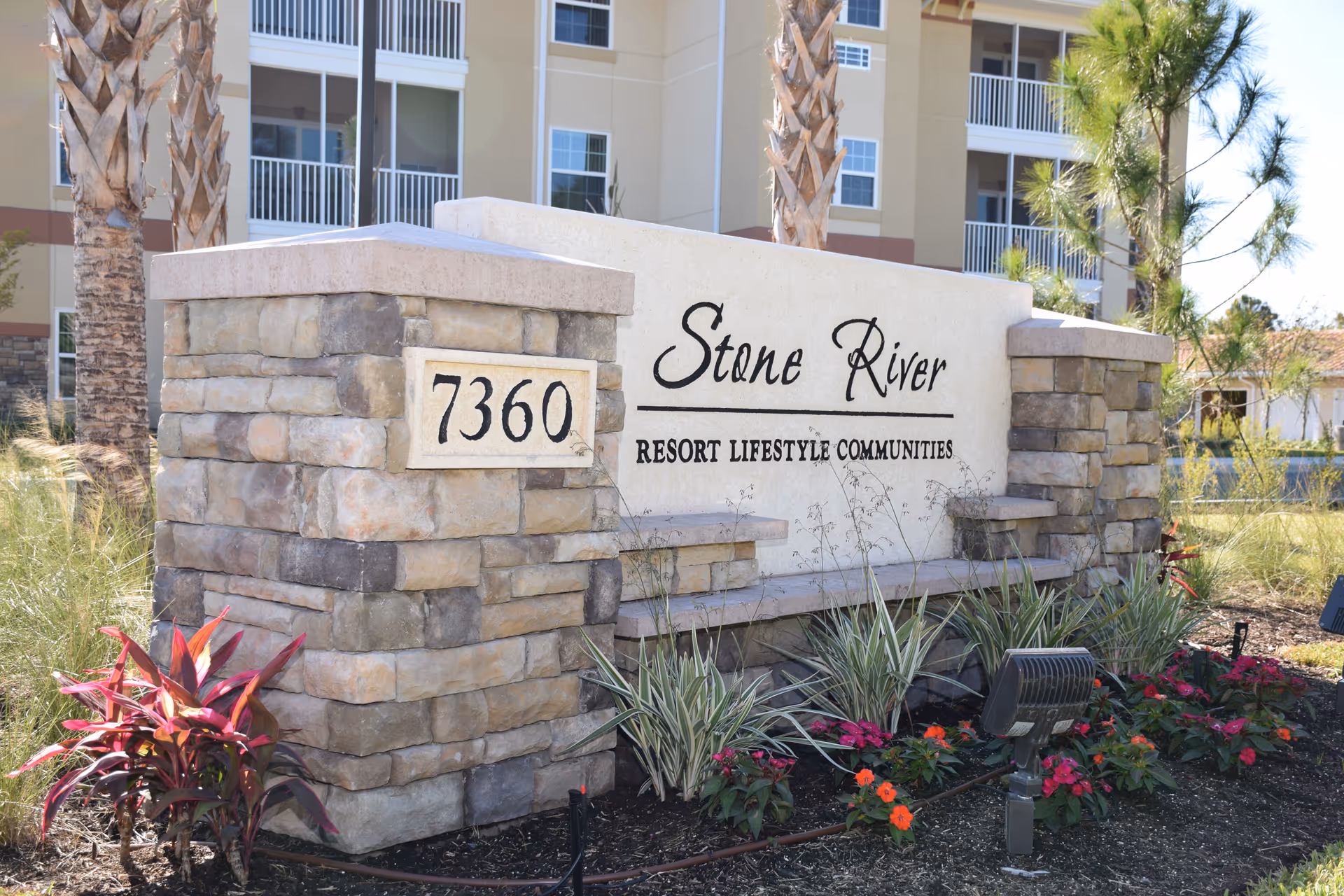 Stone River Resort Lifestyle Communities entrance sign made of stone and concrete with the address number 7360, surrounded by landscaping with plants and flowers, and a multi-story building in the background.