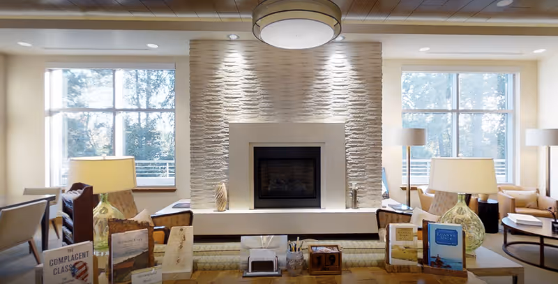 Sunlit common lounge with a central stone fireplace flanked by large windows, seating, lamps, and a table with books and decor.