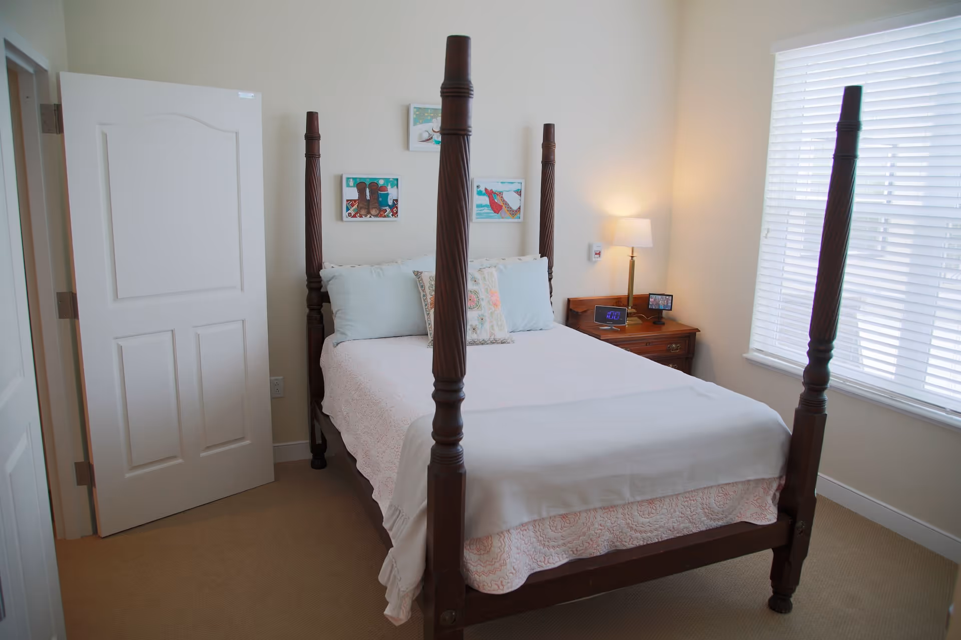 A bedroom with a four-poster wooden bed made with white and light blue bedding. There are three small framed artworks on the wall above the bed. A wooden nightstand with a lamp, digital clock, and a small picture frame is positioned next to the bed. A window with white blinds is on the right side, and a white door is open on the left side of the room.