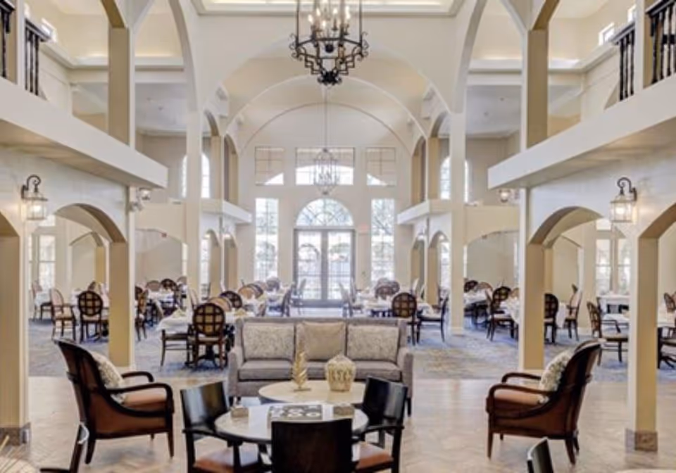 Spacious two-story communal dining and lounge area with high arched ceilings, chandeliers, and multiple seating and dining tables.
