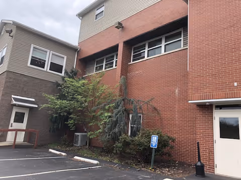Exterior view of a red-brick multi-story senior living building showing windows, a doorway, landscaping, parking spaces, and an accessible parking sign.