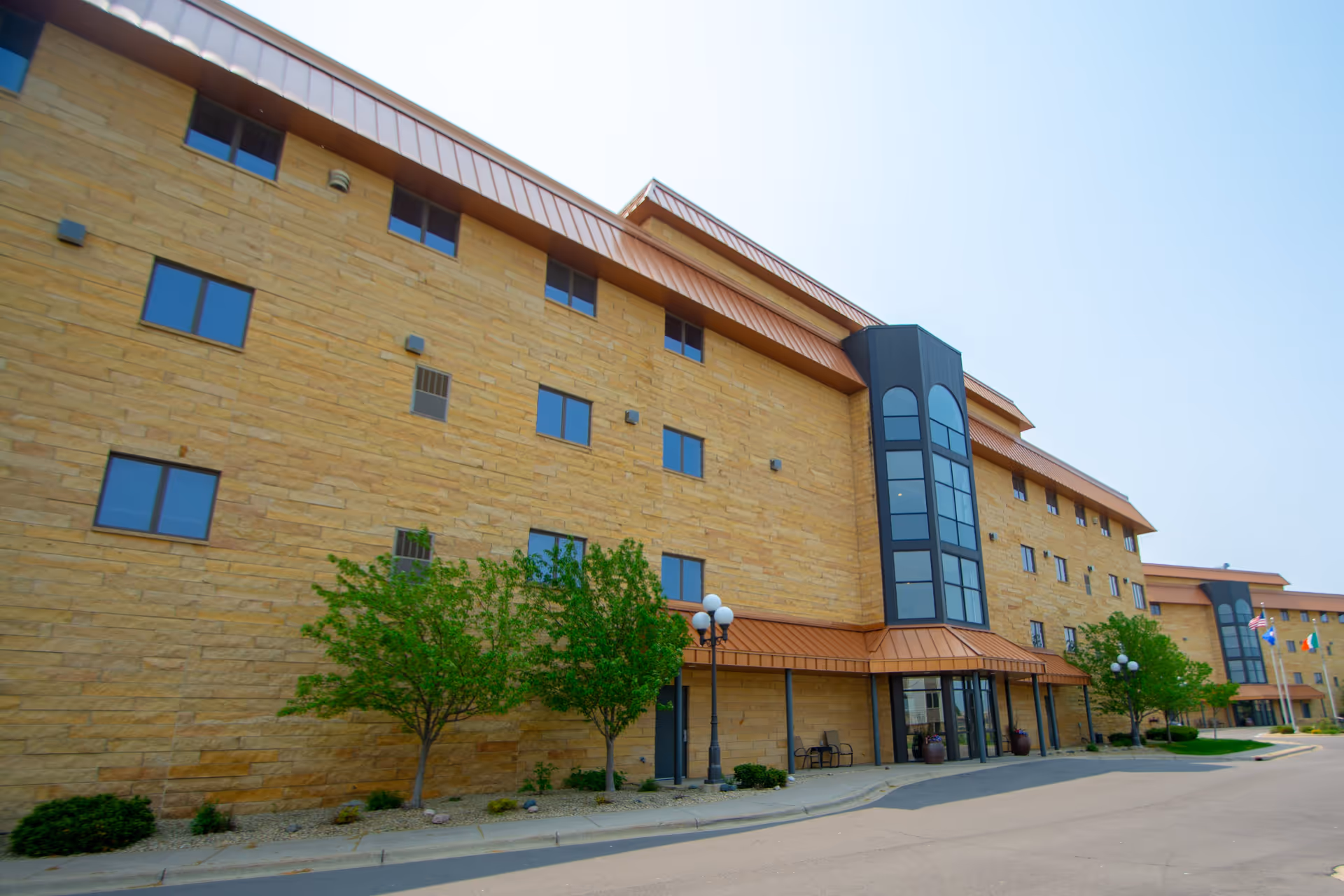 Exterior view of a multi-story senior living facility building with tan brick walls, large windows, and copper-colored roofing accents. There are small trees and shrubs planted along the sidewalk in front of the building, and a few flagpoles with flags are visible in the distance.