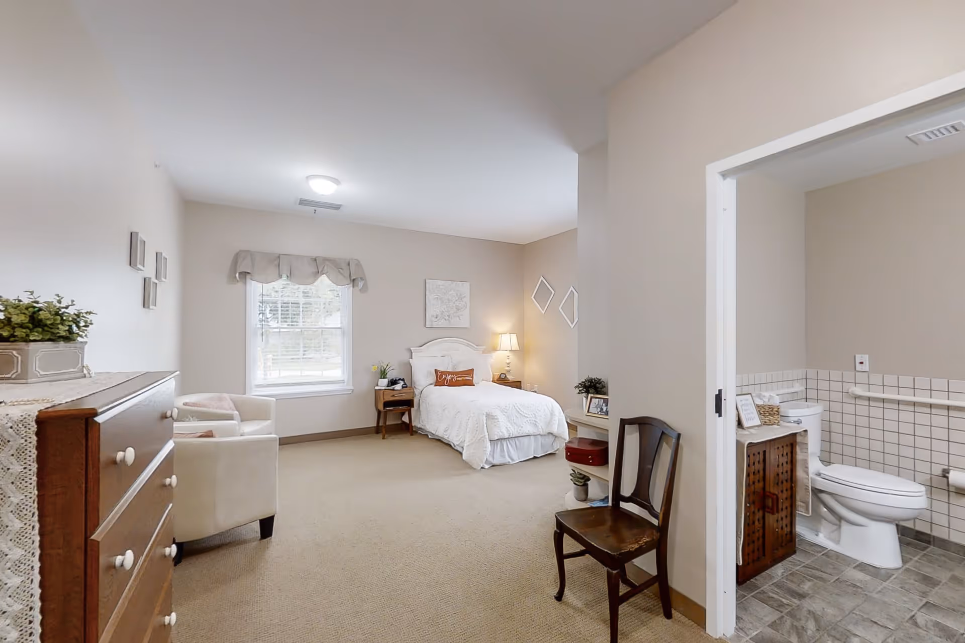 A cozy senior living bedroom with a single bed dressed in white linens, a wooden nightstand with a lamp, a window with a valance, a wooden dresser with a plant on top, a white armchair, and a wooden chair near the entrance to an adjacent bathroom. The bathroom features a toilet, tiled walls, and a wooden cabinet.
