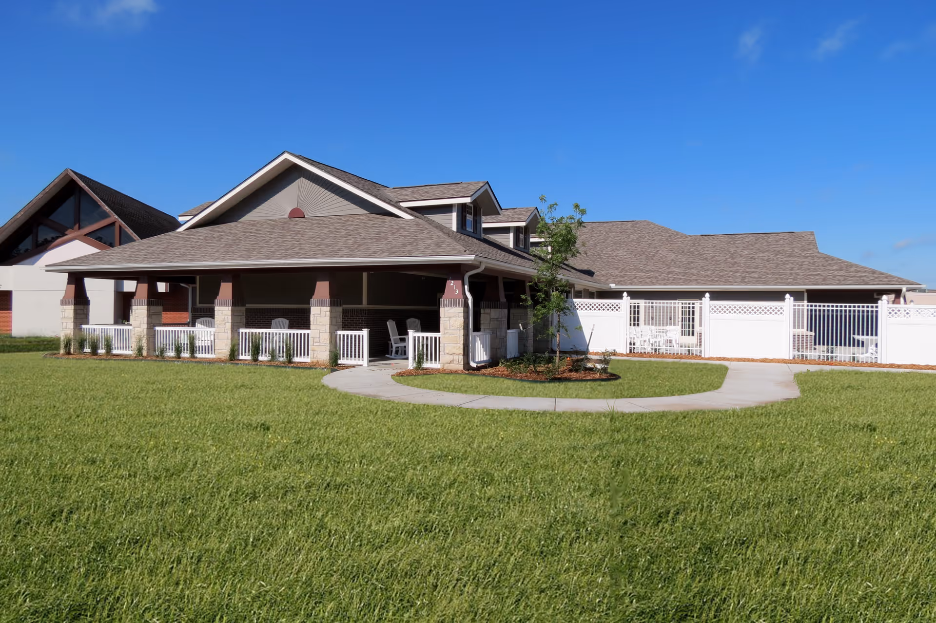 Exterior front of a single-story building with a covered porch, white railing, and a large green lawn under a clear blue sky.