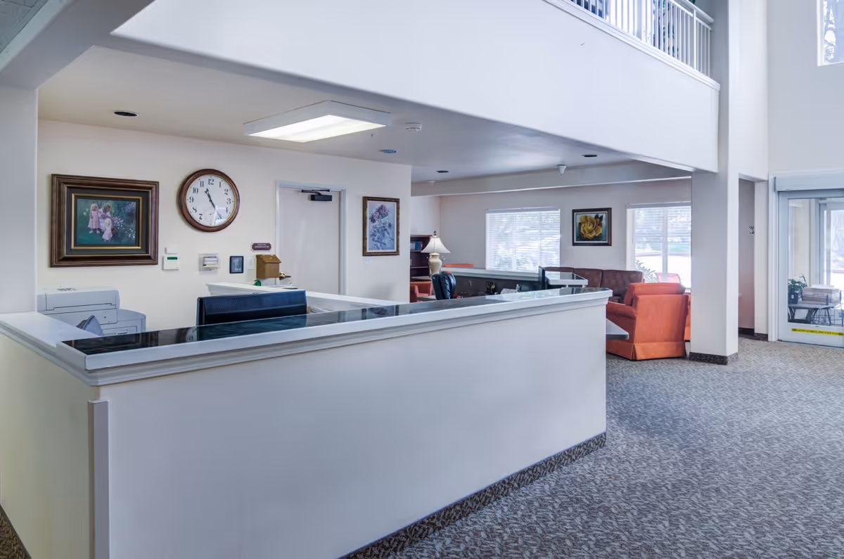 Reception area of a senior living facility with a white front desk, a clock on the wall showing 11:55, framed artwork, office equipment, and a seating area with orange chairs and a sofa near windows letting in natural light.