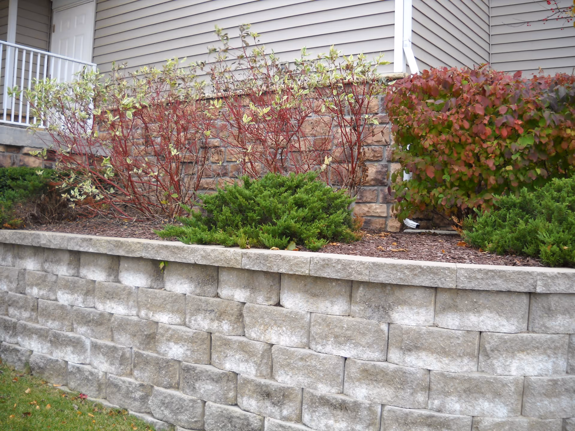 A raised garden bed with stone retaining wall in front of a building exterior. The garden contains various shrubs and plants with red and green foliage. The building has beige siding and a white door with a small porch and railing visible on the left side.