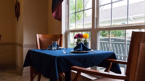 Small dining table set for two with a blue tablecloth, folded napkins, glassware and a vase of flowers beside a large window.