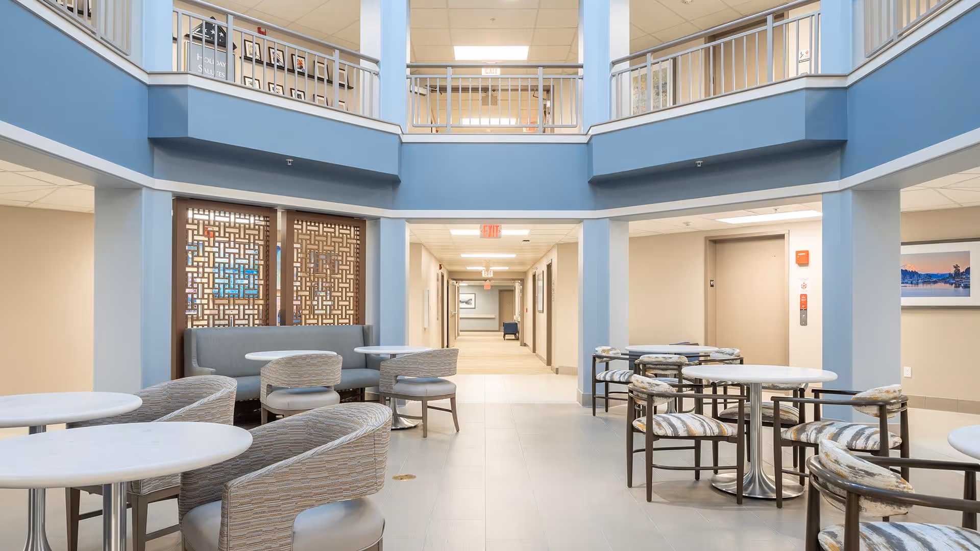 Interior view of a senior living facility common area with round tables and chairs arranged for seating. The space features a two-story ceiling with blue walls and a balcony railing on the upper level. There is a hallway leading to other rooms, an elevator, and decorative wall panels.