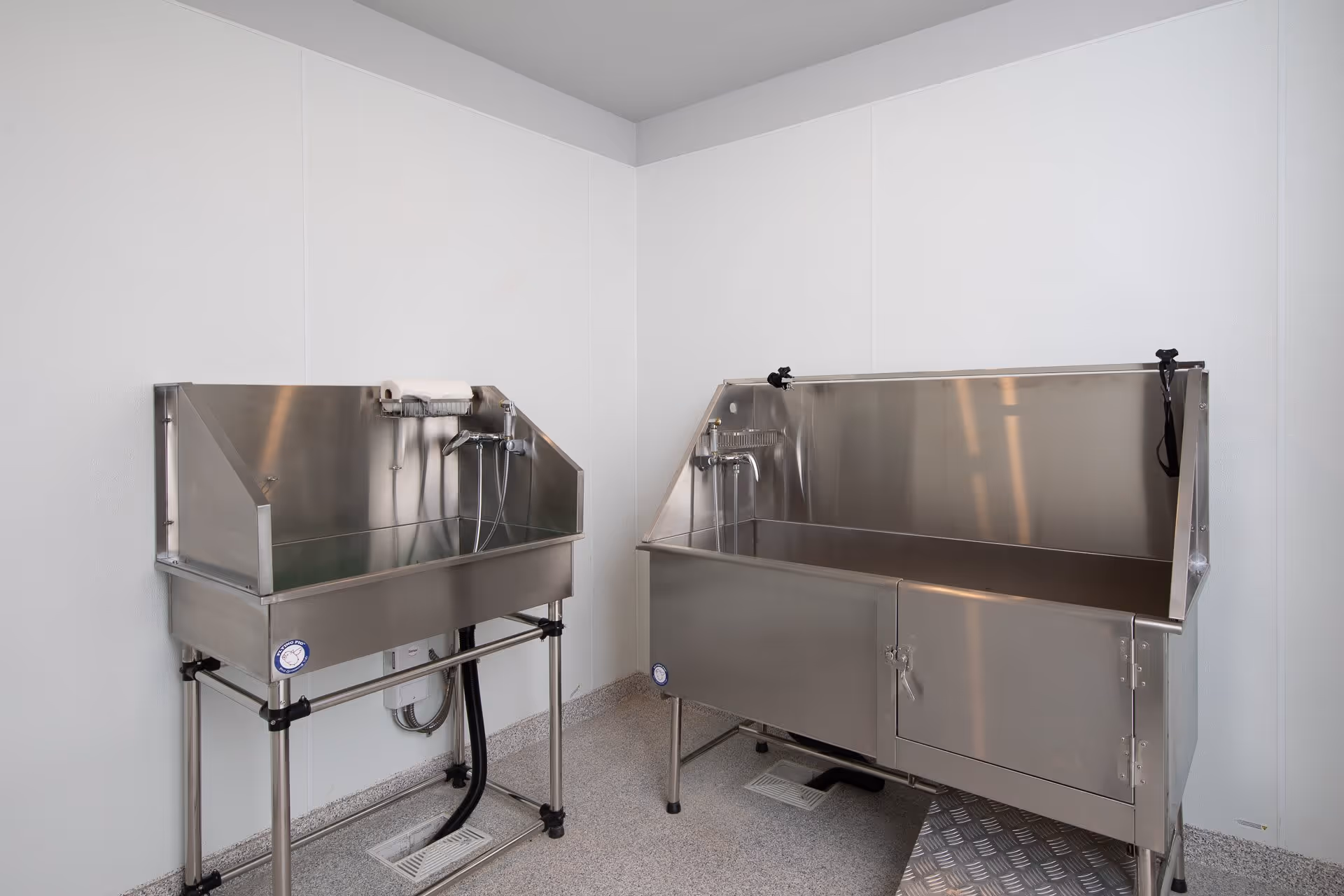 A clean room with two stainless steel bathing tubs designed for assisted bathing, each equipped with faucets and handheld shower heads, set against plain white walls and a speckled floor.