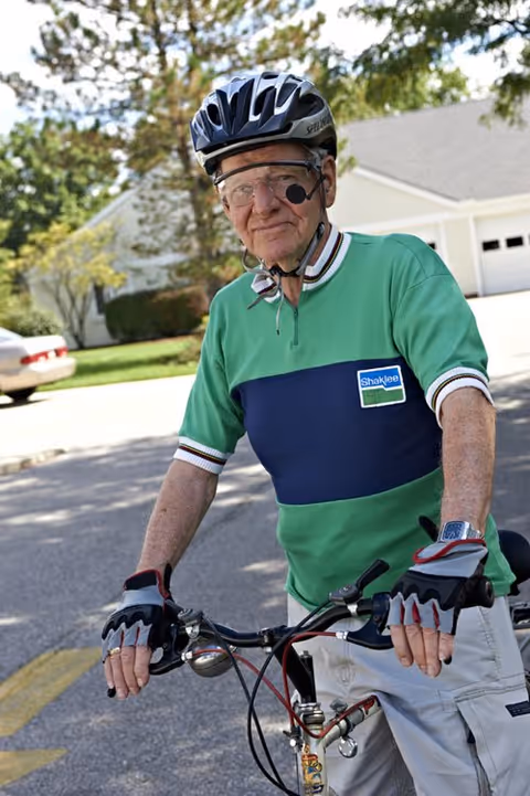 An elderly man wearing a green and navy cycling jersey, a black helmet, and protective glasses is standing outdoors with his bicycle on a residential street. There are trees, a house, and a parked car in the background.