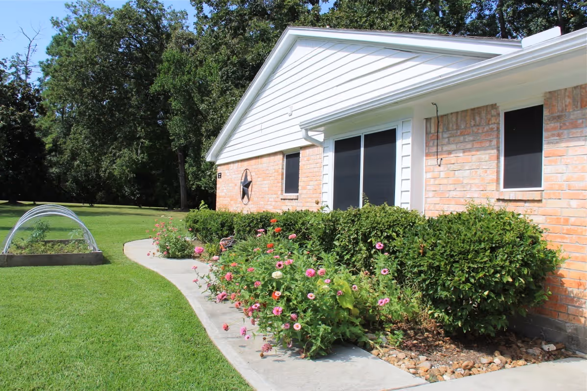 Exterior view of a brick and white siding building with a garden bed of green bushes and colorful flowers along a curved concrete walkway, surrounded by a well-maintained grassy lawn and trees in the background.