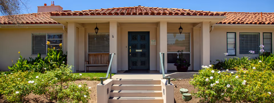 Front exterior view of a single-story building with a tiled roof, a central entrance with steps leading up to a dark green door, flanked by windows with blinds. There are plants and flowers in the garden area in front of the building under a clear blue sky.
