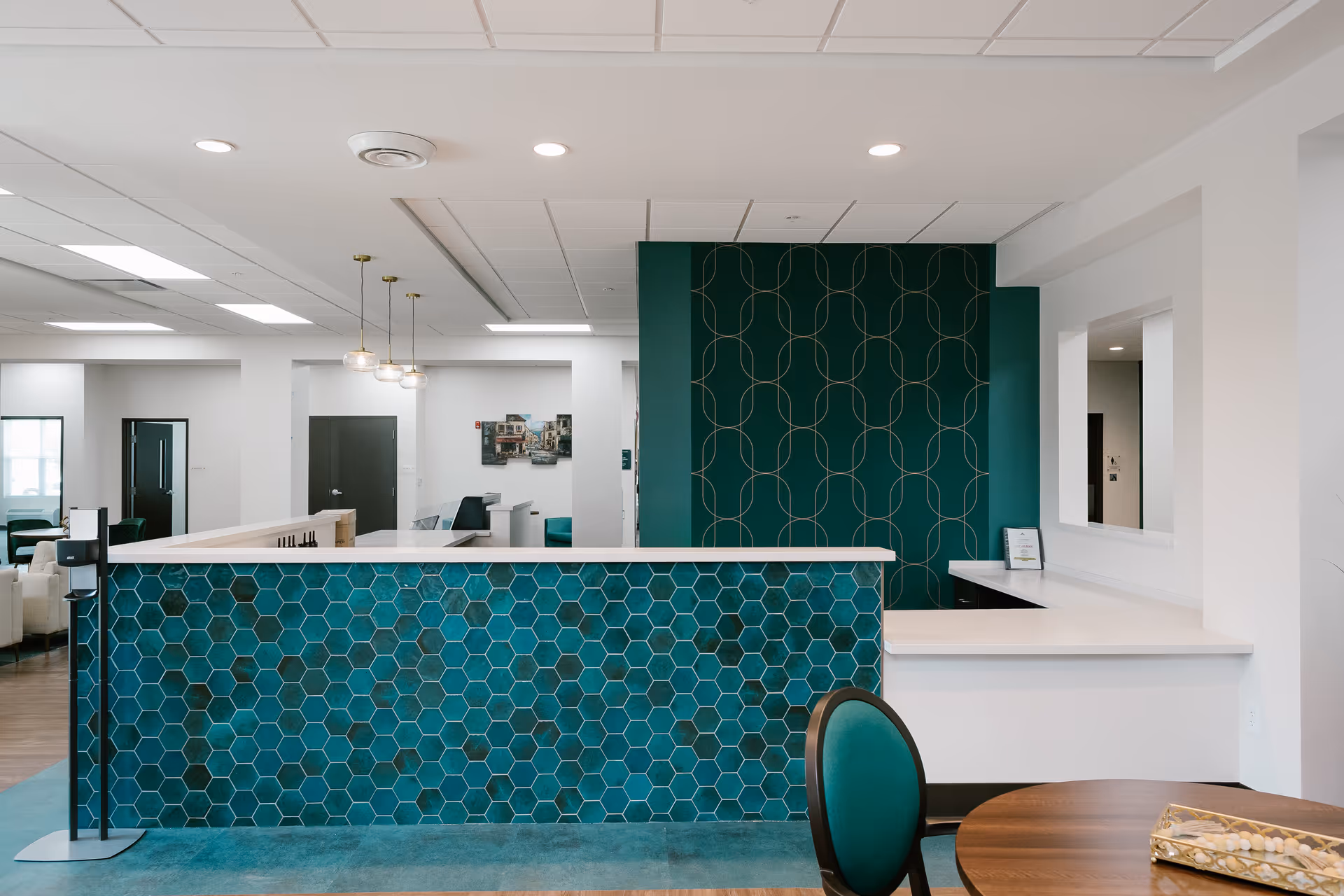 Reception area in a senior living facility with a white countertop and a front panel decorated with blue hexagonal tiles. Behind the counter is a dark green wall with a geometric pattern. The area is well-lit with ceiling lights and pendant lamps. A round wooden table with a green cushioned chair is visible in the foreground.