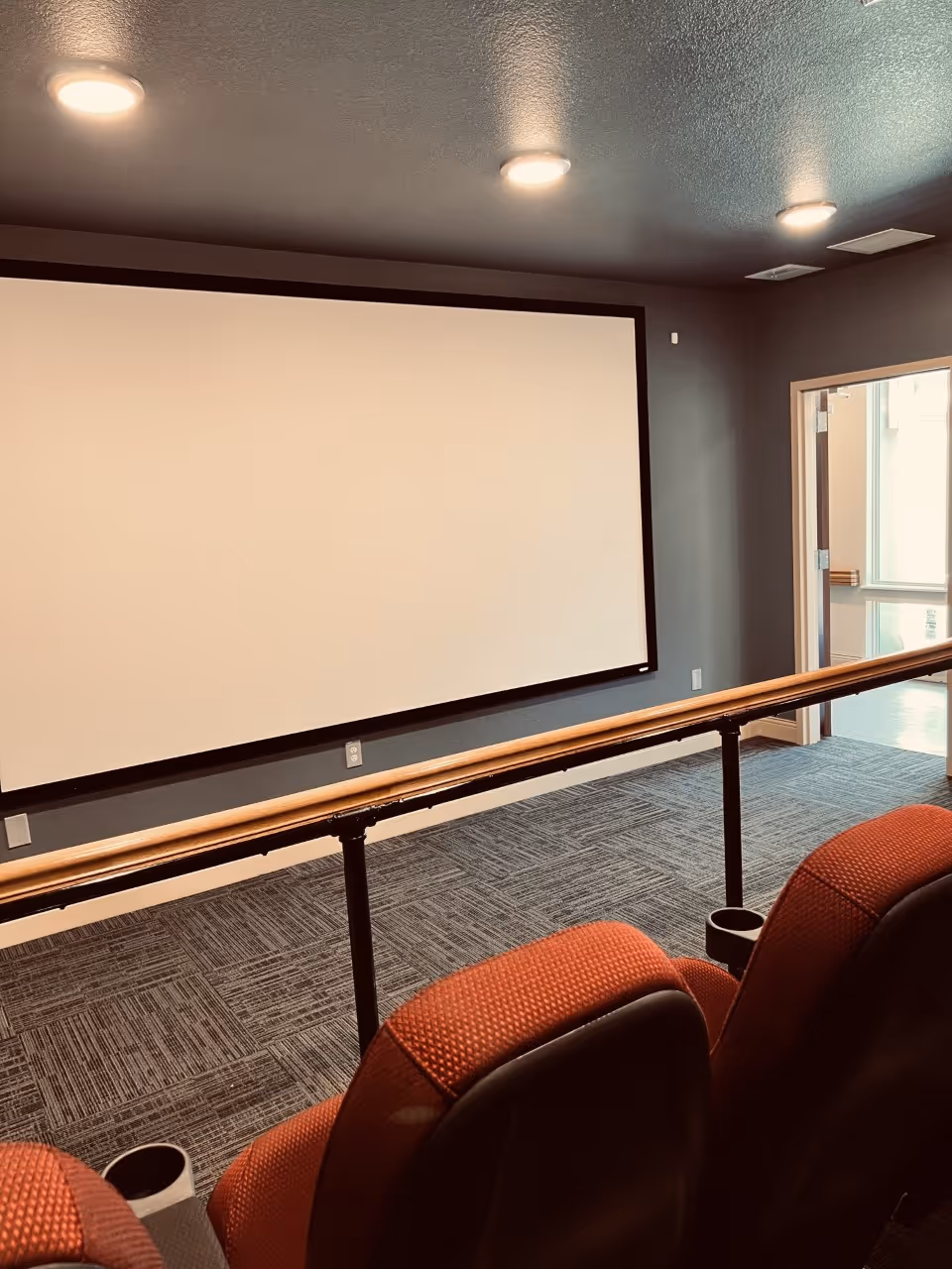 Small screening room with a large blank projection screen, red upholstered seats, and a center handrail.