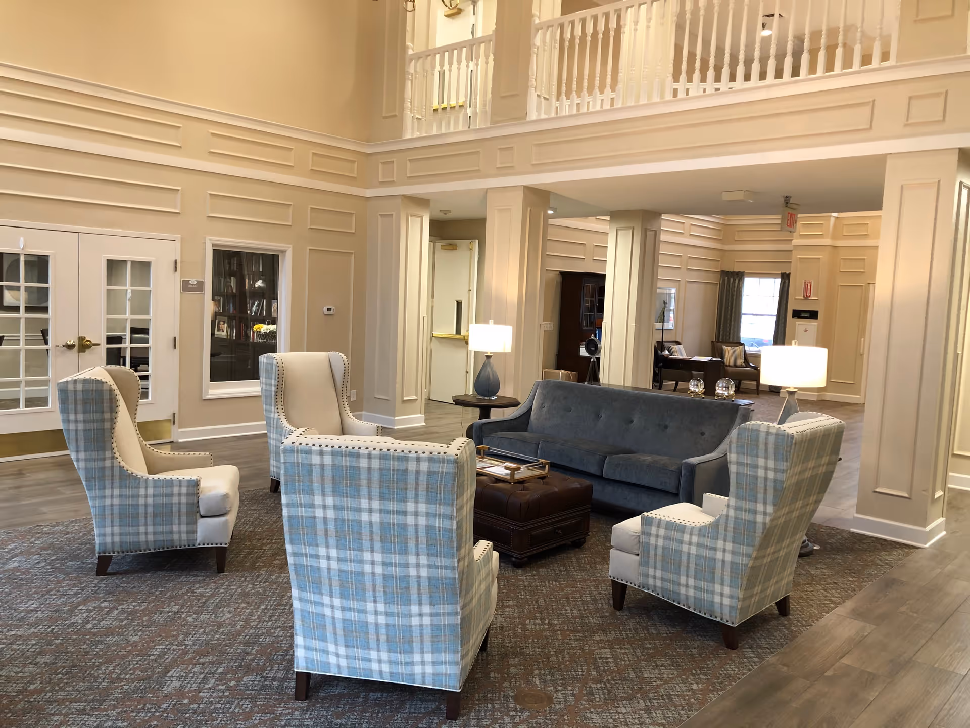 A spacious and elegant living room area in The Carriages of Rivergate featuring a blue-gray sofa, four plaid armchairs arranged around a dark brown ottoman, two side tables with lamps, and beige paneled walls with white trim. The room has a high ceiling with a balcony railing above and wood flooring partially covered by a large area rug.