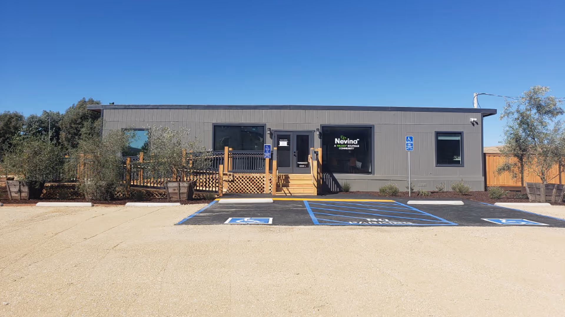 Single-story gray office building with a wooden ramp and marked handicap parking spaces in front under a clear blue sky.