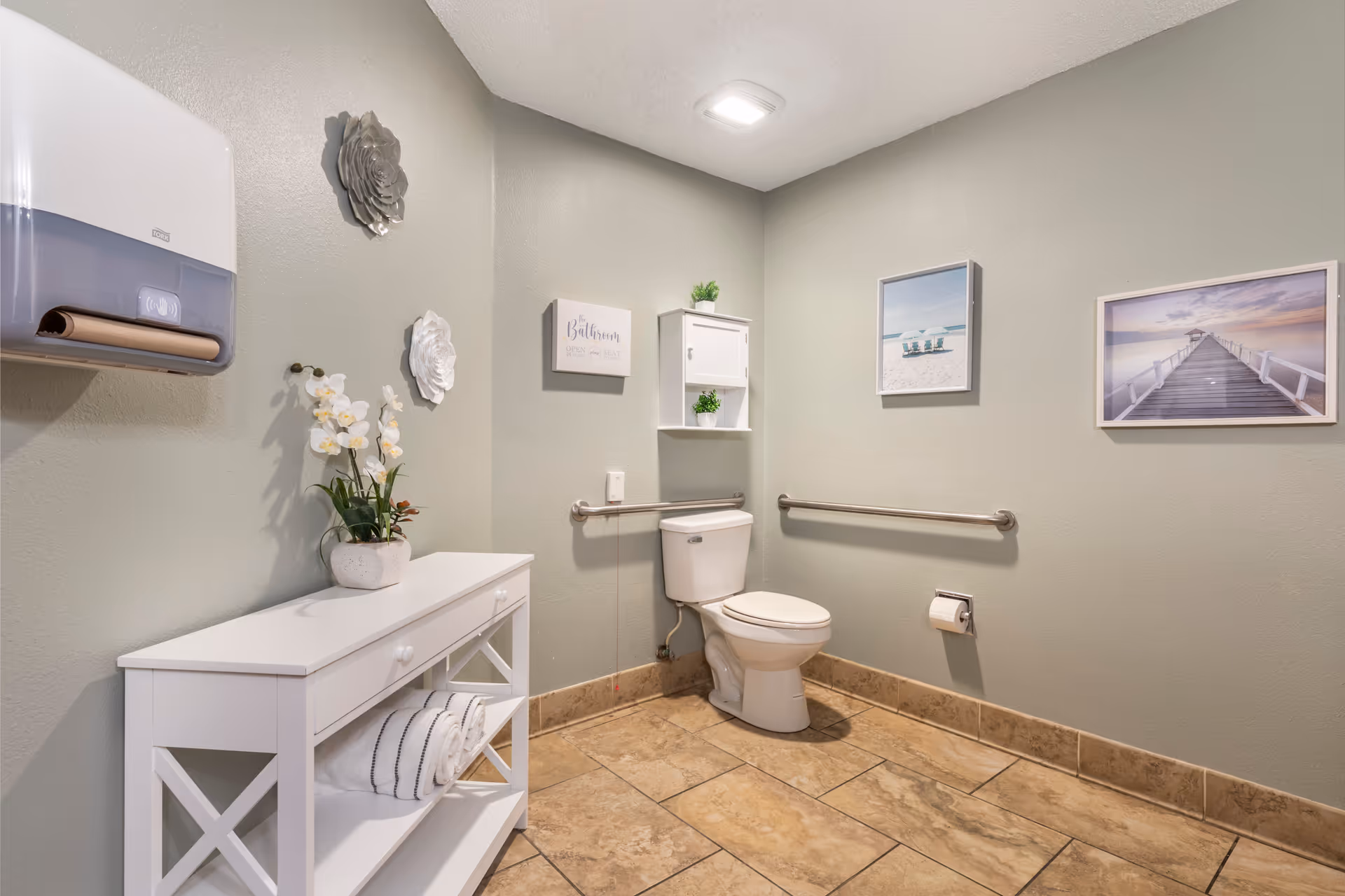 Accessible restroom with a toilet, grab bars, a white storage console holding towels and a potted plant, and framed wall art.