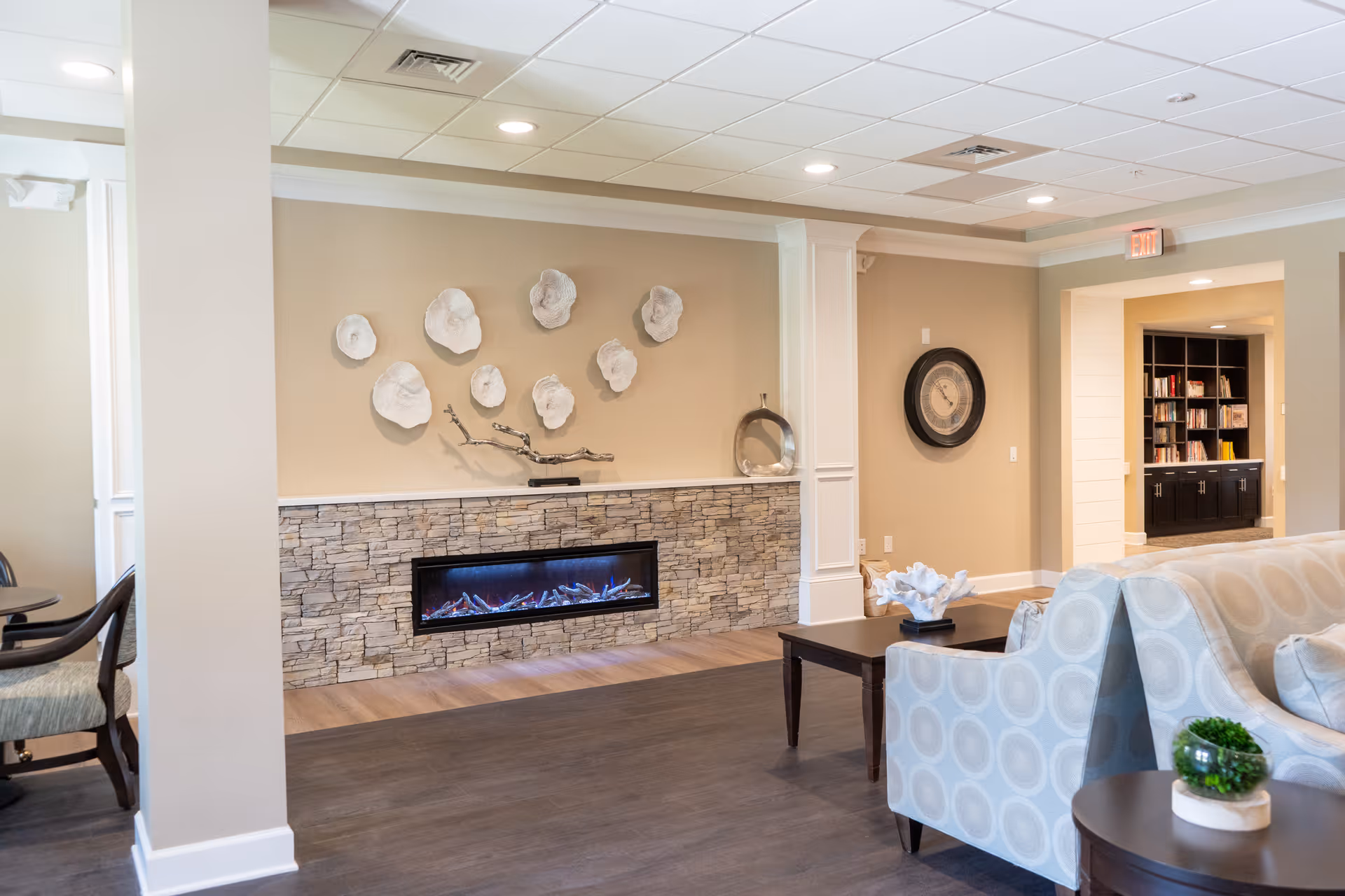 A cozy living room area with a modern electric fireplace set in a stone wall. Above the fireplace are decorative white wall sculptures resembling flowers. The room features beige walls, a large round clock, and comfortable seating with patterned upholstery. In the background, there is a bookshelf filled with books and cabinets underneath.