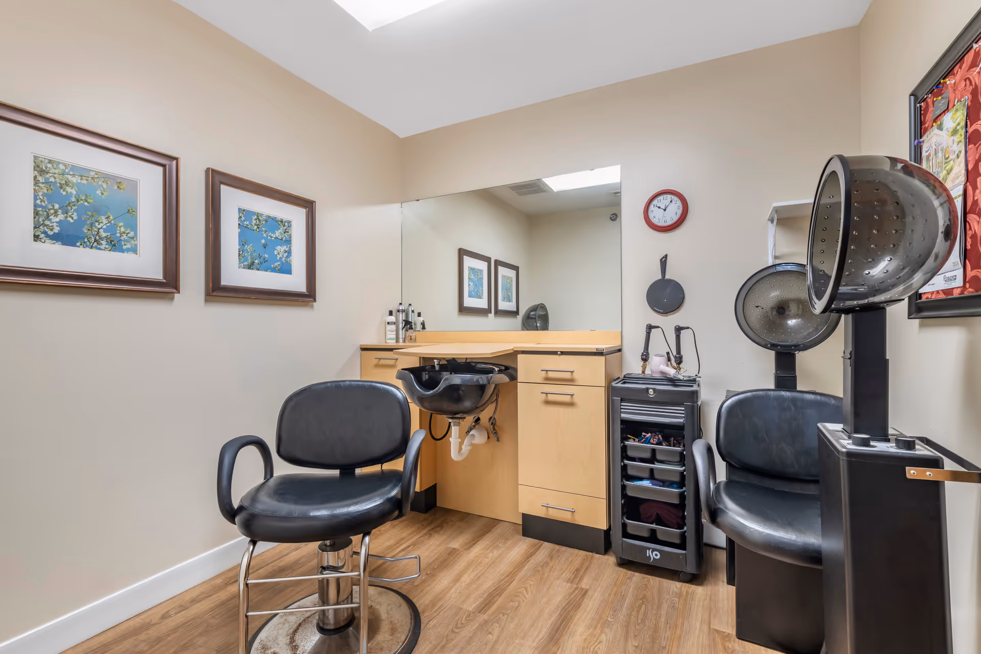 Interior of a small salon room with two black salon chairs, a hair washing sink attached to a wooden cabinet, a large mirror on the wall, two framed pictures of flowering branches, a red clock, and a hair dryer station.